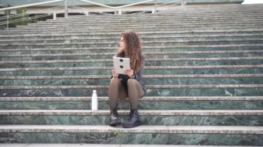 Genç bir kadın hareketli bir şehir ortamında, taş basamaklarda gelişigüzel oturur, tabletine derinden odaklanır, ki bu tablet hayatındaki yaratıcılığın ve modern teknolojinin kusursuz bir karışımını gözler önüne serer.