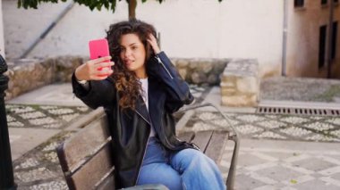 Kıvırcık saçlı genç bir kadın bir bankta oturuyor, pembe akıllı telefonuyla neşeyle selfie çekiyor, yemyeşil ve canlı çiçeklerle çevrili, doğada hoş bir anı yakalıyor.