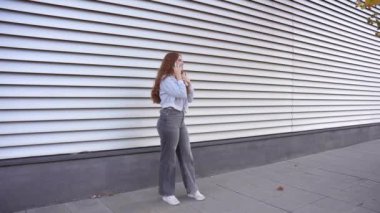 Modaya uygun genç bir kadın kendine güvenerek duruyor ve modaya uygun ama modaya uygun bir şekilde poz veriyor, çağdaş çizgili bir şehir duvarına dayanıyor, canlı, benzersiz stili ve bireyselliğini tamamen gösteriyor.