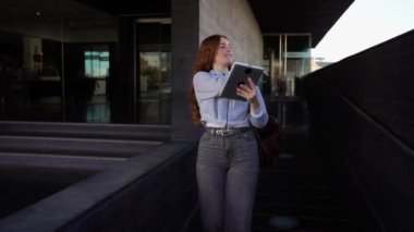 Kıvırcık kızıl saçlı genç bir kadın açık havada otururken tabletini kullanmaya kendini tamamen kaptırmış. Sıradan giyinmiş ve şık bir sırt çantası takıyor. Gerçekten güzel ve güneşli bir günün tadını çıkarıyor.