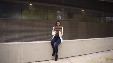 Şık bir kadın beton bir duvarda oturuyor, açık hava ortamında dizüstü bilgisayarına odaklanmış, şehir zarafetini somutlaştırıyor ve modern profesyonel değerleri yansıtan modaya uygun bir yaşam tarzı sergiliyor.