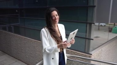 Kendine güvenen, şık beyaz bir ceket giymiş bir kadın tabletine dalmış, fırsatlarla dolu canlı bir şehir ortamında modern ve teknolojik bir yaşam tarzını örnek alıyor.