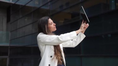 Güzel binalarla dolu bir şehir ortamında, şık genç bir kadın tabletiyle bir selfie çekerek eğlenceli bir anı yakalıyor, modern teknolojiyi ve şehir hayatının özünü gözler önüne seriyor.