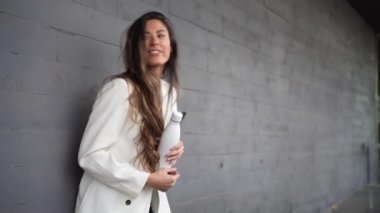 Uzun saçlı neşeli bir kadın, neşeli beyaz bir su şişesini şakacı bir şekilde elinde tutan, gri bir duvara karşı duruyor, neşe saçıyor ve enerji saçıyor.