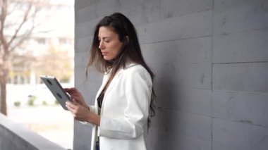 Şık bir spor ceket giyen kendine güvenen bir kadın tabletinde bir makale okur, şık gri bir duvara yaslanır, modernlik ve profesyonelliği somutlaştırırken bilgiye olan aşkını gösterir.