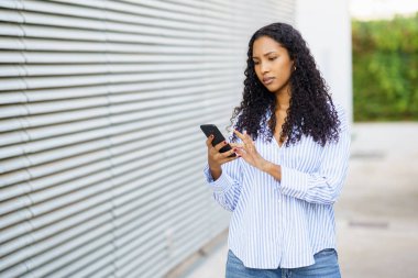 Şık çizgili gömlek ve rahat kot giyen genç bir kadın açık havada, akıllı telefonuna dikkatle bakıyor, tamamen odaklanmış ve okuduğu ya da gördüğü şeyle meşgul.