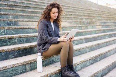 Zıplayan kıvırcık saçlı genç bir kadın, açık hava basamaklarında rahat bir şekilde oturuyor, tabletle meşgul, yanında bir su şişesi oturuyor, rahat ve modern bir yaşam tarzını güzel bir şekilde somutlaştırıyor.