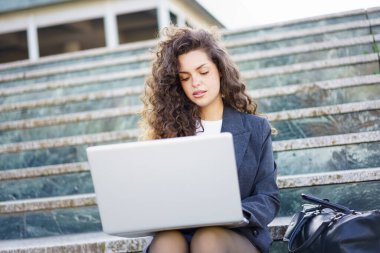 Güzel kıvırcık saçları olan genç bir kadın, merdivenlerde rahatça oturuyor. Laptopuyla yoğun bir şekilde çalışıyor. Doğa ile çevrili modern ve şık bir açık hava ortamında, gün ışığının tadını çıkarıyor.
