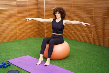 Modern bir spor salonunda, kendini adamış bir kadın denge ve dayanıklılığını geliştirmek için dengeli bir top ile pratik yapar, genel olarak formda ve sağlıklı yaşam tarzına önemli katkı sağlar.