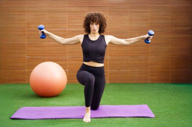 Kendini adamış bir kadın güç eğitimi için dambıllar kullanır, üst vücut gücü, çekirdek dengesi ve genel sağlık ve vücut sağlığını destekleyen eksiksiz bir egzersiz için uygun forma odaklanır.
