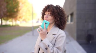 Güzel kıvırcık saçlı neşeli bir kadın, dışarıda durup, akıllı telefonunu kontrol ederken, şehrin canlı atmosferini içine çekerken sıcak güneşin tadını çıkarıyor.