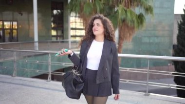 Kendine güvenen ve hırslı genç bir kadın zarif, özel dikim bir iş elbisesi içinde elinde akıllı bir telefonla fevkalade modern mimari bir arkaplan karşısında duruyor.