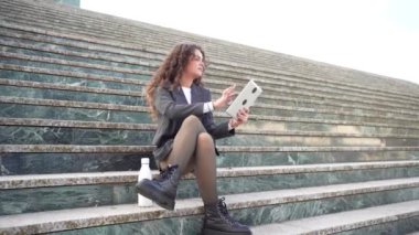 Şık bir spor ceket ve modaya uygun bir tayt giyen genç bir kadın açık hava basamaklarında oturuyor, tabletine odaklanıyor, modern iş eğilimlerini ileri teknoloji ile harmanlıyor, yaşam tarzı ve profesyonelliğini gösteriyor.