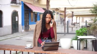 Canlı bir açık hava kafede, genç bir kadın telefonuyla coşkuyla konuşurken dizüstü bilgisayarını ustalıkla kullanıyor, kentsel yaşamın canlı özünü ve modern bağlantıyı somutlaştırıyor.