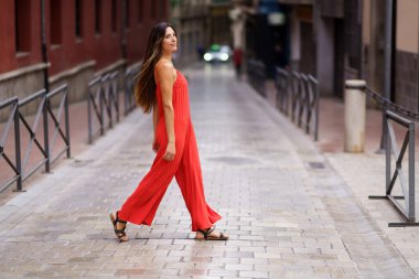 Canlı kırmızı kıyafetli kendine güvenen bir kadın huzurlu bir caddede zarif bir şekilde yürüyor, moda ve doğal güzelliğin birlikteliğini somutlaştırıyor, neşeli tavırlarıyla herkesi büyülüyor.