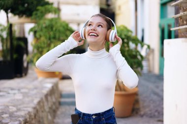 Şehir ortamında, şık kulaklıklı neşeli genç bir kadın açık havada neşe içinde müzik dinliyor, mutluluk, rahatlama ve müziğin getirdiği heyecan verici özgürlük.