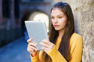 Parlak sarı kazaklı genç bir kadın tabletini canlı şehir ortamında yıpranmış taş bir duvara karşı kullanıyor. Dijital çağımızdaki modern bağlantıyı vurguluyor.