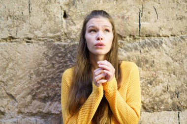 Parlak sarı bir kazak giyen genç bir kadın düşünceli bir şekilde yukarı bakar. Yüzünde merak ve umut karışımı bir ifade vardır. Dokulu taş bir duvara yaslanırken düşlerinde kaybolur.