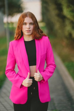 Tarz sahibi genç bir kadın canlı pembe bir ceket ve modaya uygun siyah bir hasat bluzuyla poz veriyor. Açık hava güzelliğiyle çevrelenmiş, modern moda yeteneğini ön plana çıkarıyor.