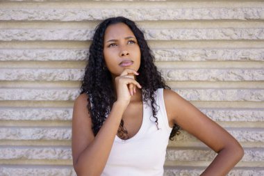 Güzel kıvırcık saçlı genç bir kadın, dokulu bir duvara yaslanırken derin düşüncelere dalmış, mükemmel bir şekilde bir yansıma ve iç gözlem anı yakalıyor, kendi derin dünyasında kayboluyor.