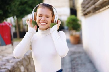 Neşeli genç bir kadın dışarıda müzik dinliyor, şık kulaklıklar takıyor. Neşeli ifadesi mutluluk ve özgürlüğü yansıtıyor.