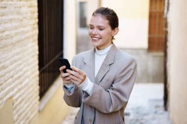 Şık giyinmiş neşeli genç bir kadın, büyüleyici bir sokakta yürürken akıllı telefonuyla meşgul oluyor.