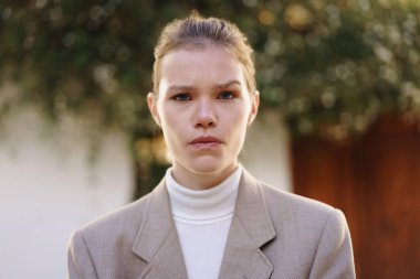 Şık bir balıkçı yaka kazak ile birlikte şık bir ceket giyen dengeli genç bir kadın, güzel bir doğa manzarası ile çevrili iken kendine güvensizlik havası yayar.