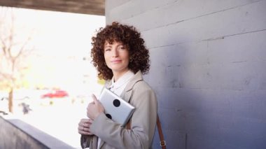 Kıvırcık saçlı, kendine güvenen profesyonel bir kadın elinde önemli belgelerden oluşan kalın bir klasör olan modern bir duvara karşı duruyor. Şehir ortamında kararlılık ve zarafet yayıyor.