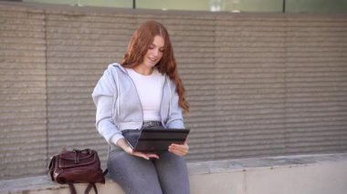 Canlı yeşillikle dolu manzaralı bir parkta, kızıl saçlı genç bir kadın tabletine dalmış, şık giyinmiş, şık tasarımcı çantasının yanında sıcak güneşin tadını çıkarıyor, rahatlamak için mükemmel.