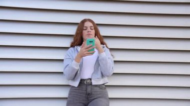 Canlı, kıvırcık kızıl saçlı genç bir kadın şık, modern bir duvara yaslanıp rahat ve modaya uygun yaşam tarzını sergilerken akıllı telefon cihazına odaklanıyor.