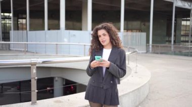 Uzun, kıvırcık saçlı şık bir kadın, elinde akıllı telefonuyla modern bir binanın yanında duruyor. Yoldan geçenleri büyüleyen şehir atmosferini büyüleyen bir cazibe saçıyor.