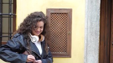 Güzel kıvırcık saçlı, şık deri ceketli, kulaklık takmış, zarif ve şık genç bir kadın çantasına dikkatlice bakıyor çarpıcı derecede canlı sarı bir duvara karşı.