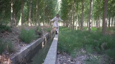 Huzurlu bir su yolunun yanında uzanan dar bir yolda dikkatle yürüyen bir insan, güzel bir şekilde yemyeşil ağaçlar ve huzurlu bir atmosfer yaratan canlı bir yeşillik ile çevrilidir.