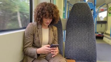 Güzel kıvırcık saçlı genç bir kadın rahat bir şekilde trende oturuyor, akıllı telefonuna dalmış, rahat atmosferini tamamlayan sakin ve sakin bir seyahat arkaplanı ile çevrelenmiş.