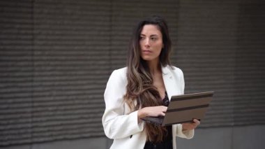 Uzun saçlı, şık elbisesiyle şık görünen profesyonel bir kadın tabletine dalmış bir şekilde dışarıda duruyor ve modern teknolojinin kurumsal bir ortama entegrasyonunu vurguluyor.