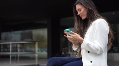 Şık, şık bir ortamda, odaklanmış ve nişanlı genç bir kadın açık havada rahat bir şekilde oturuyor, akıllı telefonuyla etkileşime geçiyor, günümüz dünyasında modern iletişimin özünü mükemmel bir şekilde sergiliyor.