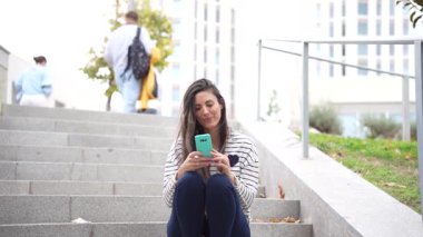 Genç bir kadın gün boyunca canlı bir şehir ortamının keyfini çıkarırken akıllı telefonuyla mutlu mesut taş basamaklarda oturuyor.