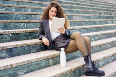 Genç bir kadın açık hava merdivenlerinde oturuyor, büyüleyici bir kitaba dalmış, şık giyinmiş, elinde modaya uygun bir su şişesi tutuyor, modaya uygun bir yaşam tarzını somutlaştırıyor.