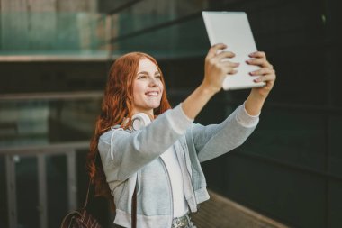 Neşeli ve hayat dolu kızıl saçlı genç bir kadın tabletini kullanarak bir selfie çekiyor. Bu sırada çeşitli modern mimarilerle dolu canlı bir şehir arkaplanı sergiliyor.