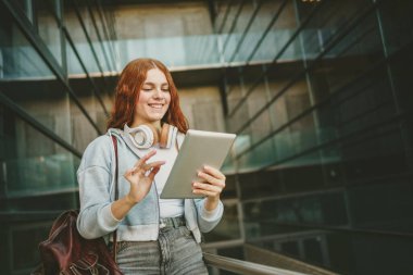 Parlak kızıl saçlı canlı bir genç kadın şık bir tablet tutuyor ve modaya uygun kulaklıklar takıyor, hareketli bir şehir ortamında rahat ve şevkle modern bir yaşam tarzını somutlaştırıyor.