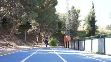 Ağaçlarla çevrili mavi bir pistte koşan bayan sporcu güneşli bir açık hava sporunun tadını çıkarıyor.