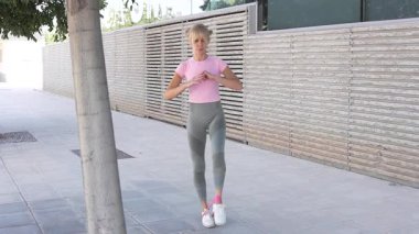 Pembe bluzlu ve yeşil tozluklu fit bir kadın, bir direniş bandıyla açık havada egzersiz yapıyor, fiziksel zindelik ve genel olarak iyi olmak için çeşitli atletik aktiviteler sergiliyor.