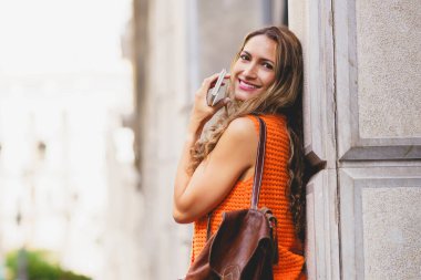 Latin bir kadın kentin dış cephesine yaslanmış elinde cep telefonu ve şehirde gülümsüyor.