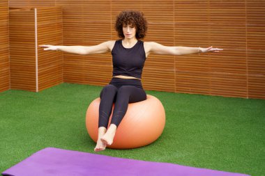 Bir kadın denge topu kullanırken etkileyici bir denge egzersizi yapar. Değişik fitness ekipmanlarıyla dolu dinamik bir spor salonunda gücünü ve istikrarını açıkça gösterir.