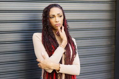 Uzun, güzel örgülü saçları olan düşünceli bir kadın, özgün stili ve kendini ifade ediş tarzını büyüleyici bir şekilde sergileyerek, zengin desenli bir arka plana doğru poz veriyor.