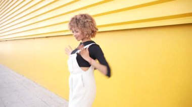 Zarif genç bir kadın, enerjik bir sarı duvarın önünde dans ederken zarafet ve akıcılığını sergiliyor. Neşeli moda anlayışını ve neşeli ruhunu mükemmel bir şekilde yansıtıyor.