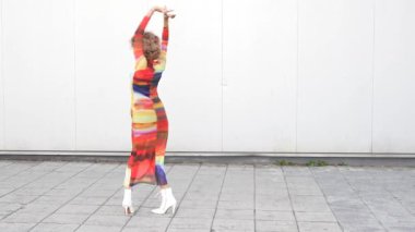 Şık bir kadın zarif, renkli ve şatafatlı bir elbiseyle dans ediyor. Moda tarzını, cazibesini arttıran minimalist beyaz bir arka plana karşı sergiliyor.