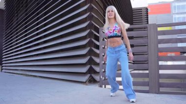 Canlı sarışın bir kadın kendine güven ve neşe saçarken çağdaş bir şehir binasının önünde mavi kot pantolon ve renkli bir bluzla enerjik bir şekilde dans ediyor. Dinamik ve şık bir ifade sergiliyor.