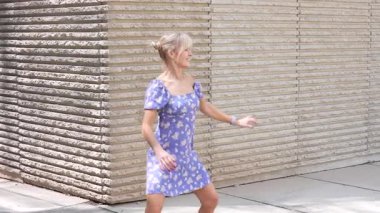 Parlak güneş ışığı altında, şık mavi çiçekli bir elbise giyen neşeli bir kadın açık havada tuğla bir duvarın yanında duruyor, modern, kentsel ortamında kaygısız ve hareketli bir tavır sergiliyor.