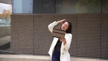 Şık giyinmiş kendine güvenen bir kadın modern tabletiyle meşgul, çağdaş bir şehir duvarına karşı zarif bir şekilde oturuyor, özgüven ve özgüven ile teknolojinin karışımını sergiliyor.